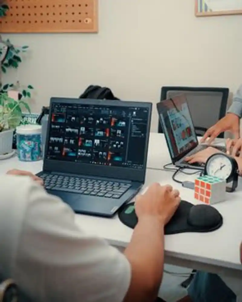 Dua orang sedang bekerja menggunakan laptop di meja kantor modern dengan kopi dan sebuah kubus Rubik.